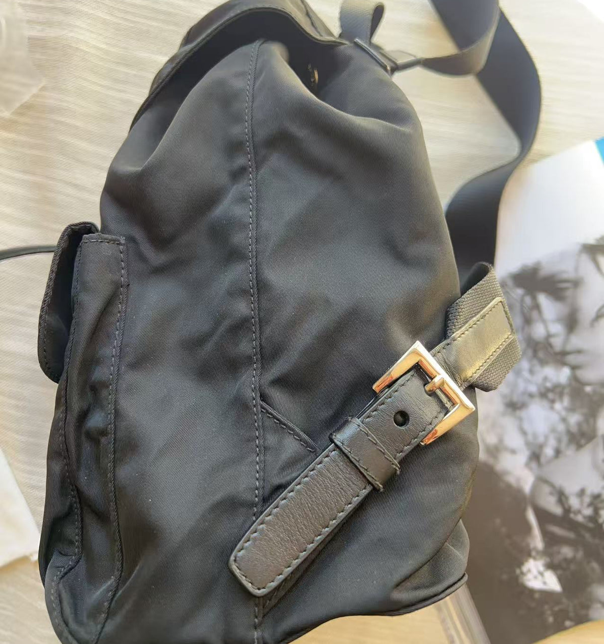 A black nylon shoulder bag with a brown leather strap and a gold-tone buckle, resting on a light wood surface next to an open magazine.