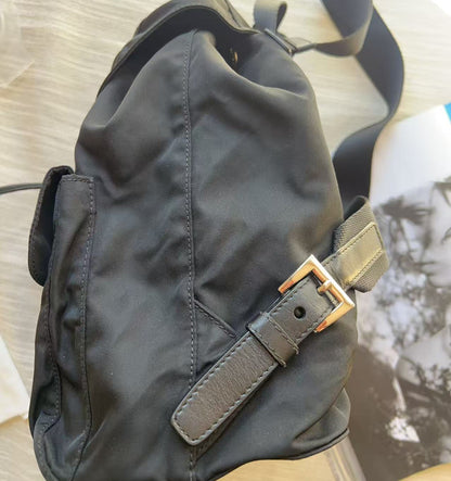 A black nylon shoulder bag with a brown leather strap and a gold-tone buckle, resting on a light wood surface next to an open magazine.