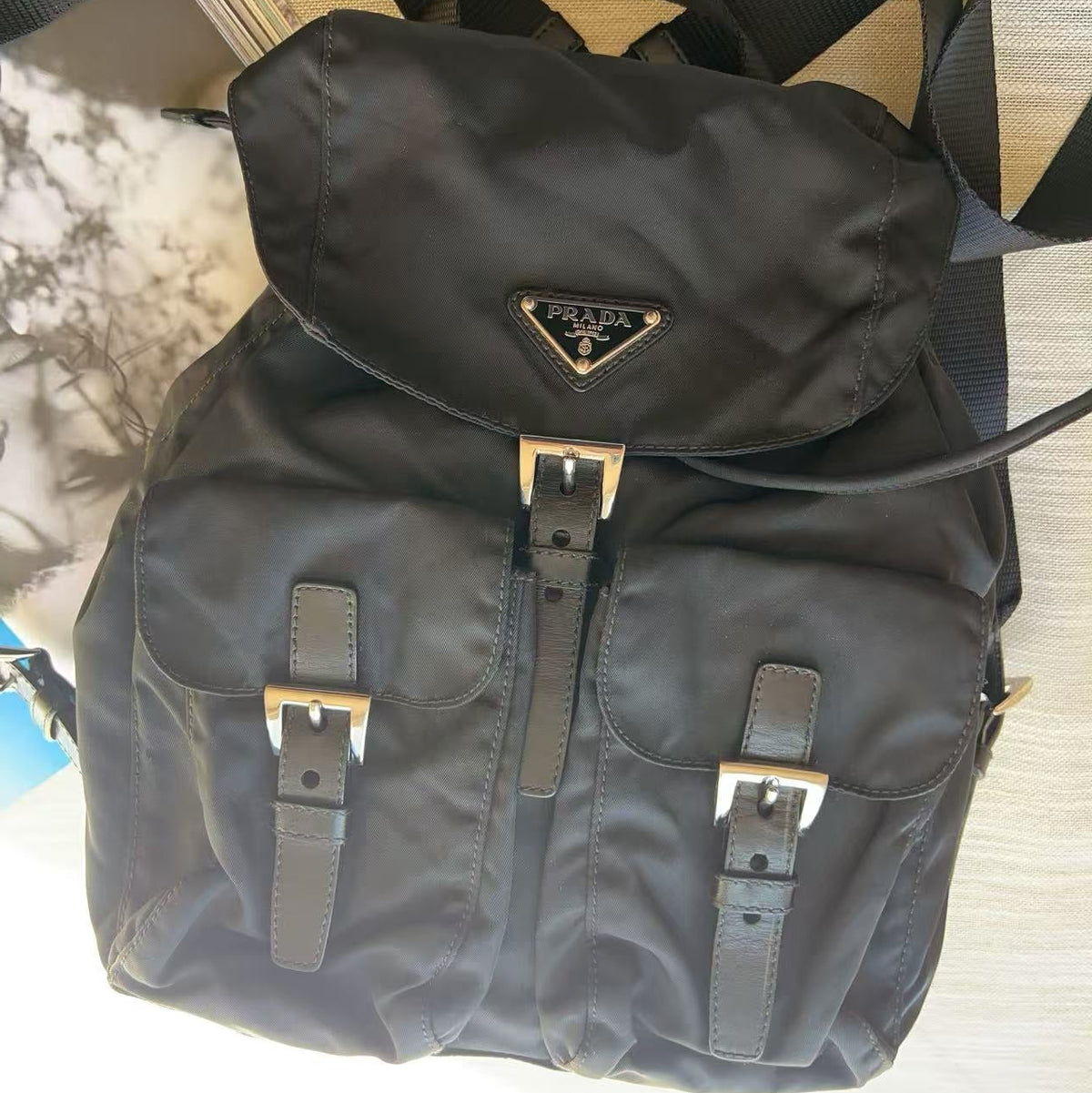 A black nylon backpack with dual front flap pockets, adjustable straps, and a small triangular metal logo plaque on the front, photographed on a light surface.