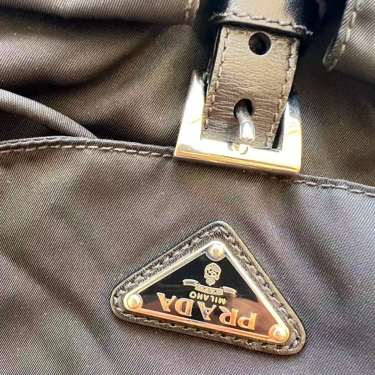 Close-up of a Prada nylon bag with a triangular metal logo plaque and a leather strap with a silver buckle.