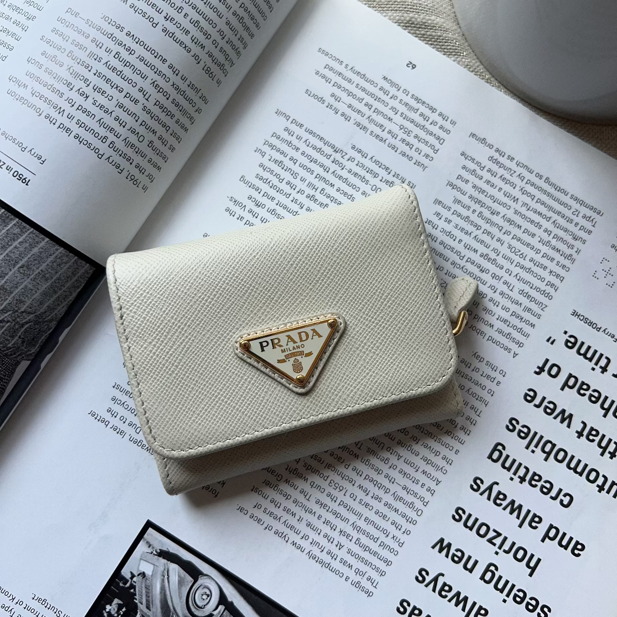 A cream-colored Prada wallet with a gold-tone triangle logo clasp, resting on an open book with a white ceramic cup in the background.
