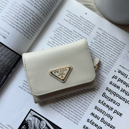 A cream-colored Prada wallet with a gold-tone triangle logo clasp, resting on an open book with a white ceramic cup in the background.