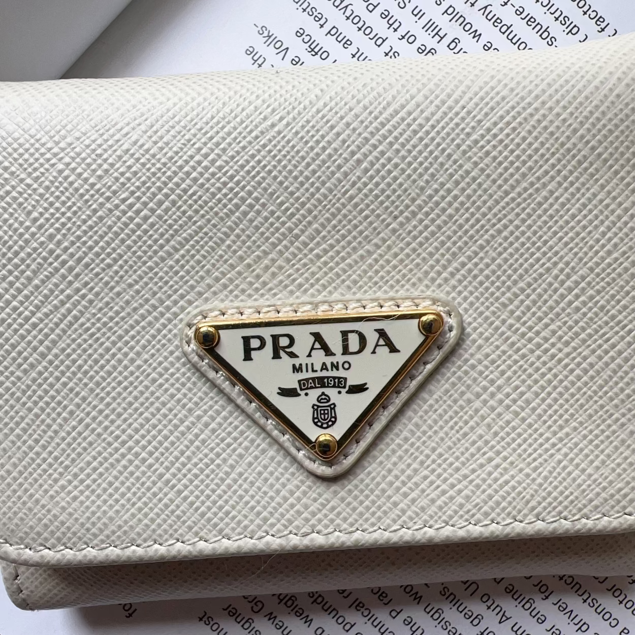 A close-up of a Prada wallet in textured off-white leather, featuring the iconic gold-trimmed triangular Prada logo plaque on the front, resting on printed book pages.