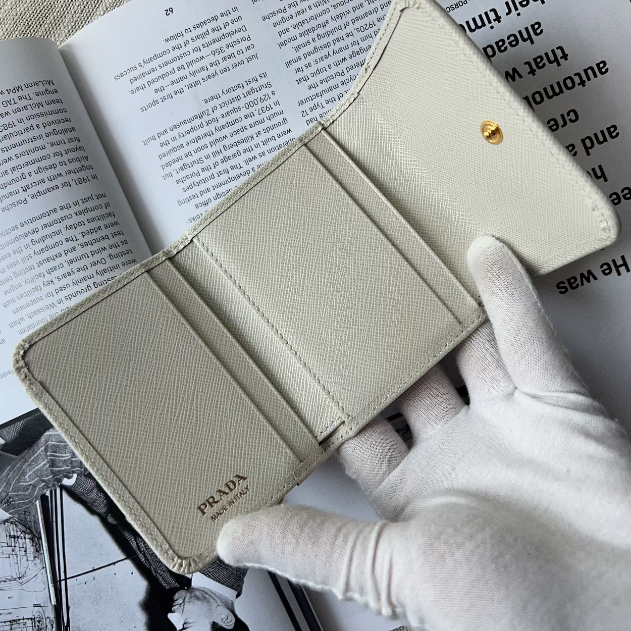 A Prada wallet in light beige textured leather, held open by a hand wearing a white glove over an open book.