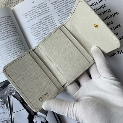 A Prada wallet in light beige textured leather, held open by a hand wearing a white glove over an open book.