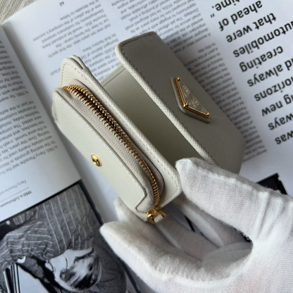 A small cream-colored leather zip-around wallet with gold hardware is held by a white-gloved hand over an open book.