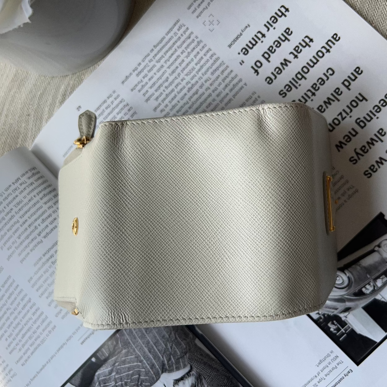 A light beige textured leather zippered pouch with gold-tone hardware, resting on an open book over a neutral fabric surface.
