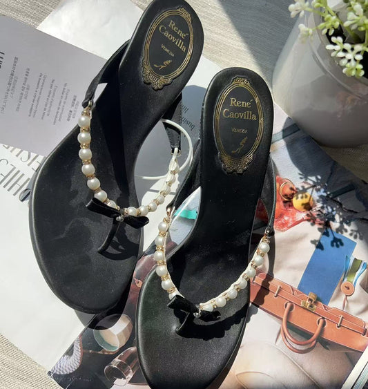 A pair of black thong sandals with pearl straps and small black bow details, displayed on top of a magazine page with a small potted plant in the background.