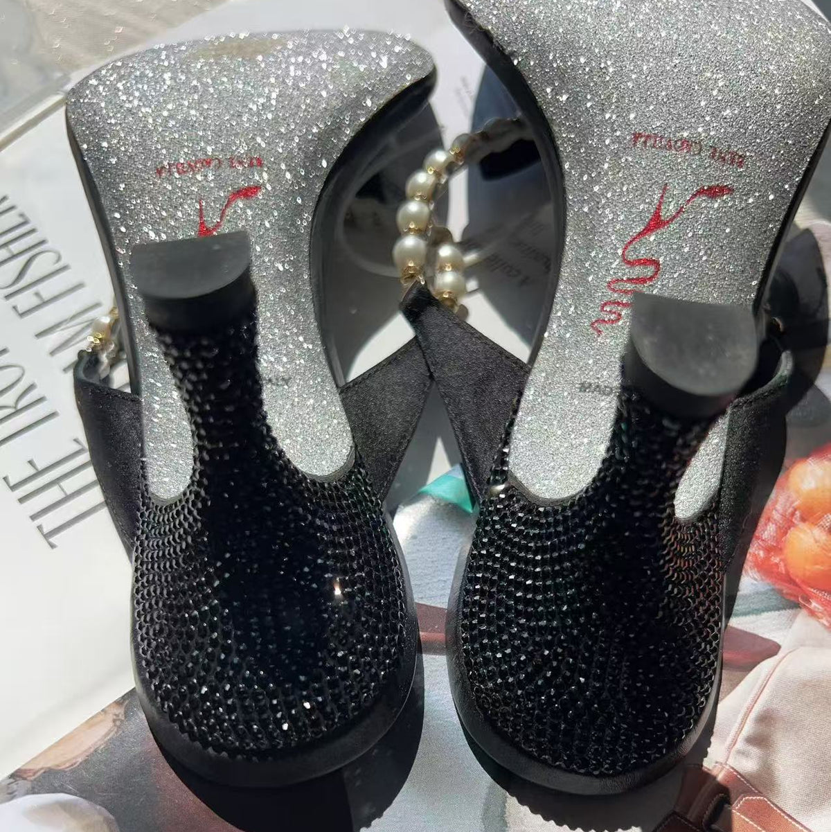 A pair of black and silver high-heeled mule sandals covered in rhinestones, shown from the top on a styled flat-lay background with a magazine and leather accessories.