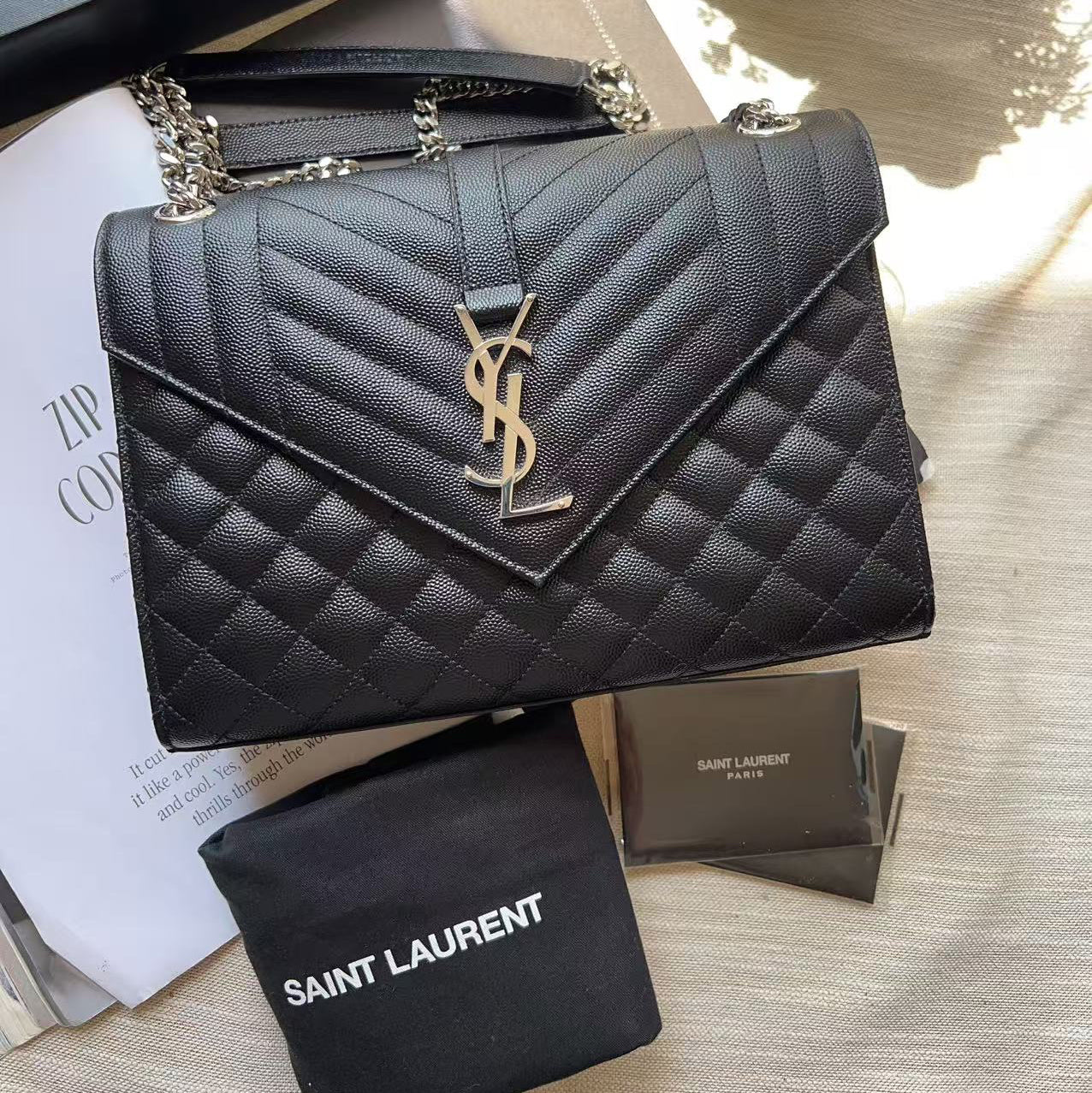 A black quilted Saint Laurent shoulder bag with a gold YSL logo, displayed with its black dust bag and packaging on a light fabric surface.