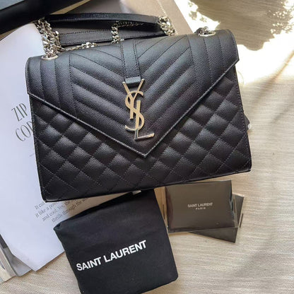 A black quilted Saint Laurent shoulder bag with a gold YSL logo, displayed with its black dust bag and packaging on a light fabric surface.