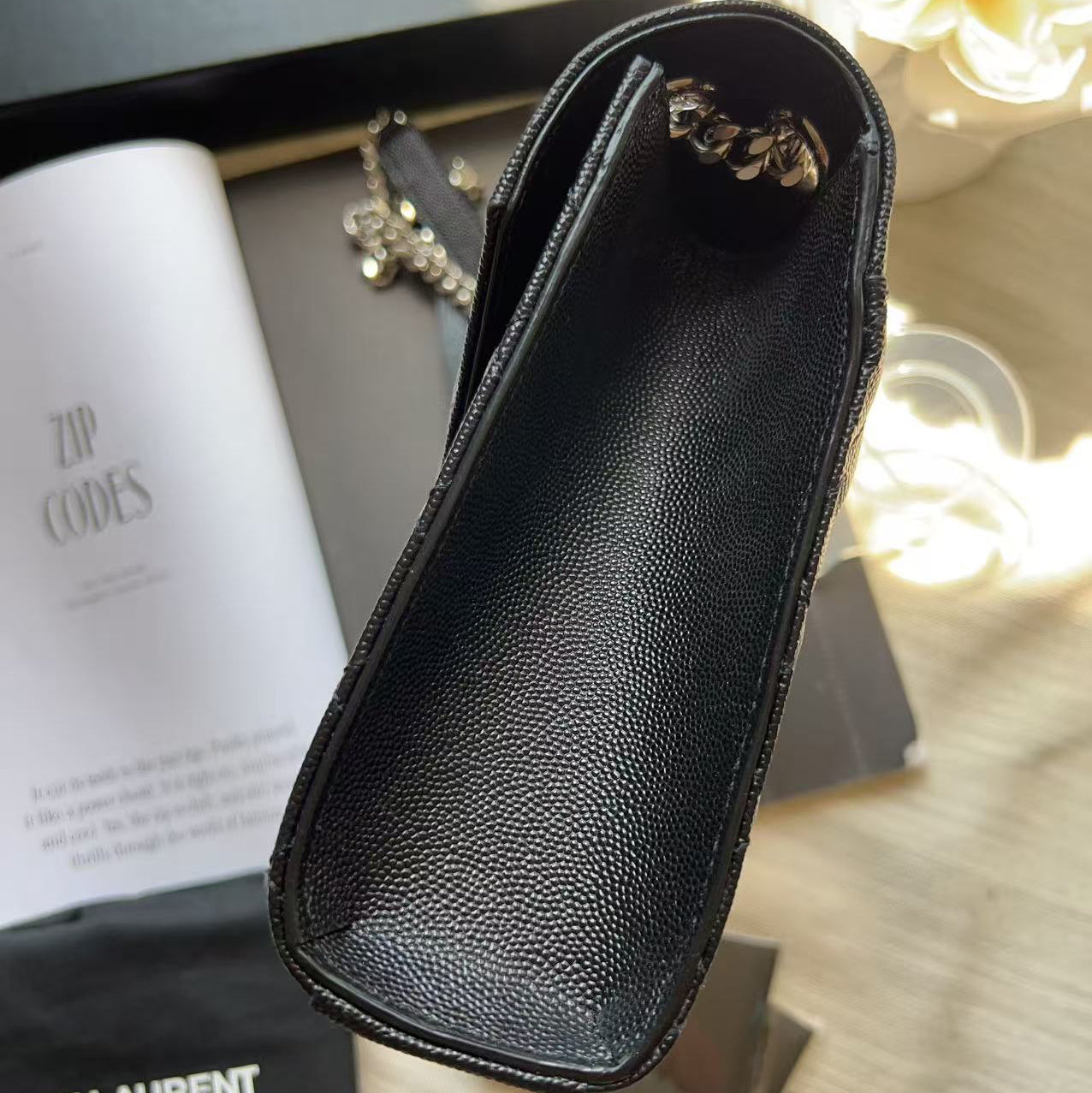 A black Saint Laurent zippered pouch with a pebbled leather texture, shown in a close-up shot with its branded dust bag, box, and manual on a wooden surface.