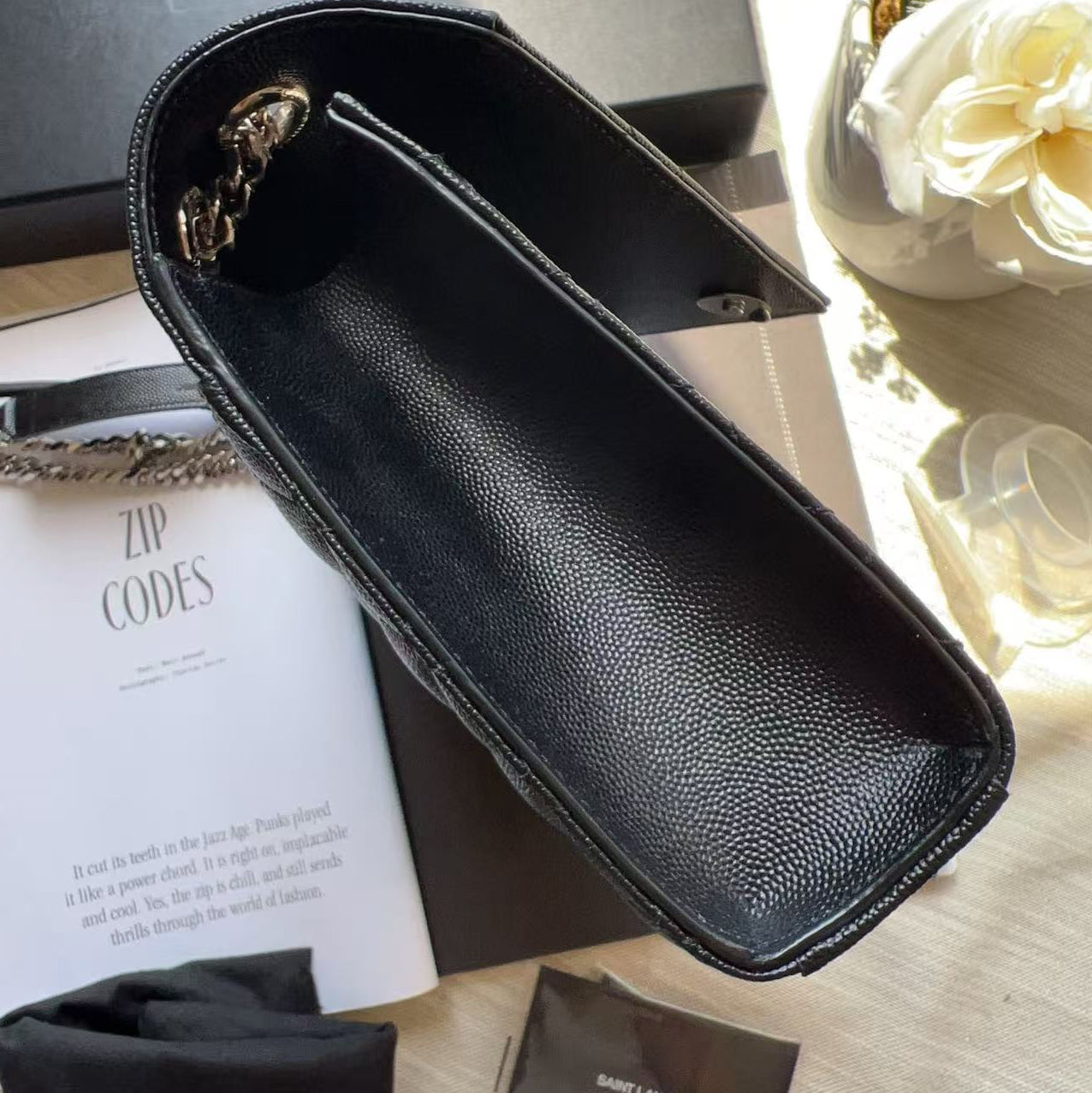 A black pebbled leather Saint Laurent pouch or organizer, shown open with a gold-tone zipper pull. The product is displayed on top of its black Saint Laurent box, an open instruction booklet, and a black dust bag on a light-colored wooden surface.