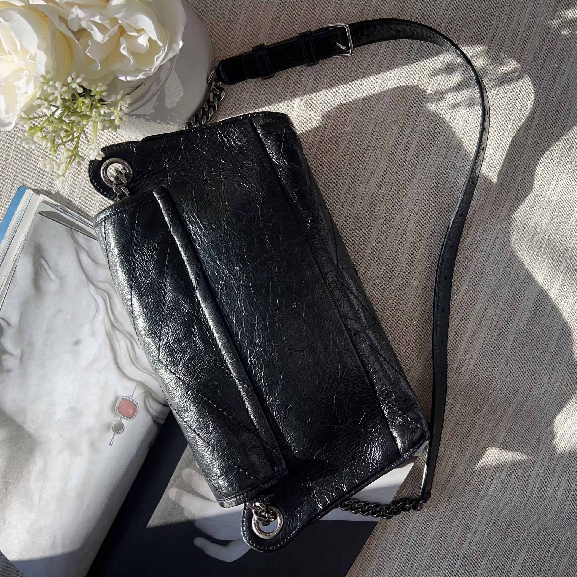 Black leather handbag with chain strap on a textured surface with flowers and magazine in the background