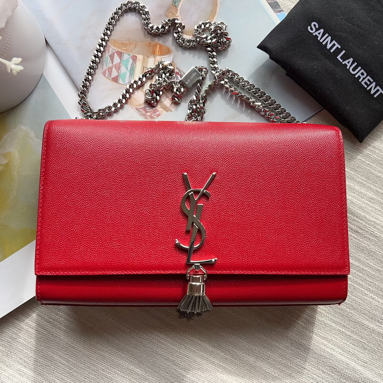 Red leather wallet with a brand logo on a textured surface with a magazine and necklace.