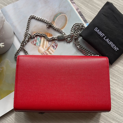 Red handbag with chain detail on a textured surface, accompanied by a black Saint Laurent pouch.