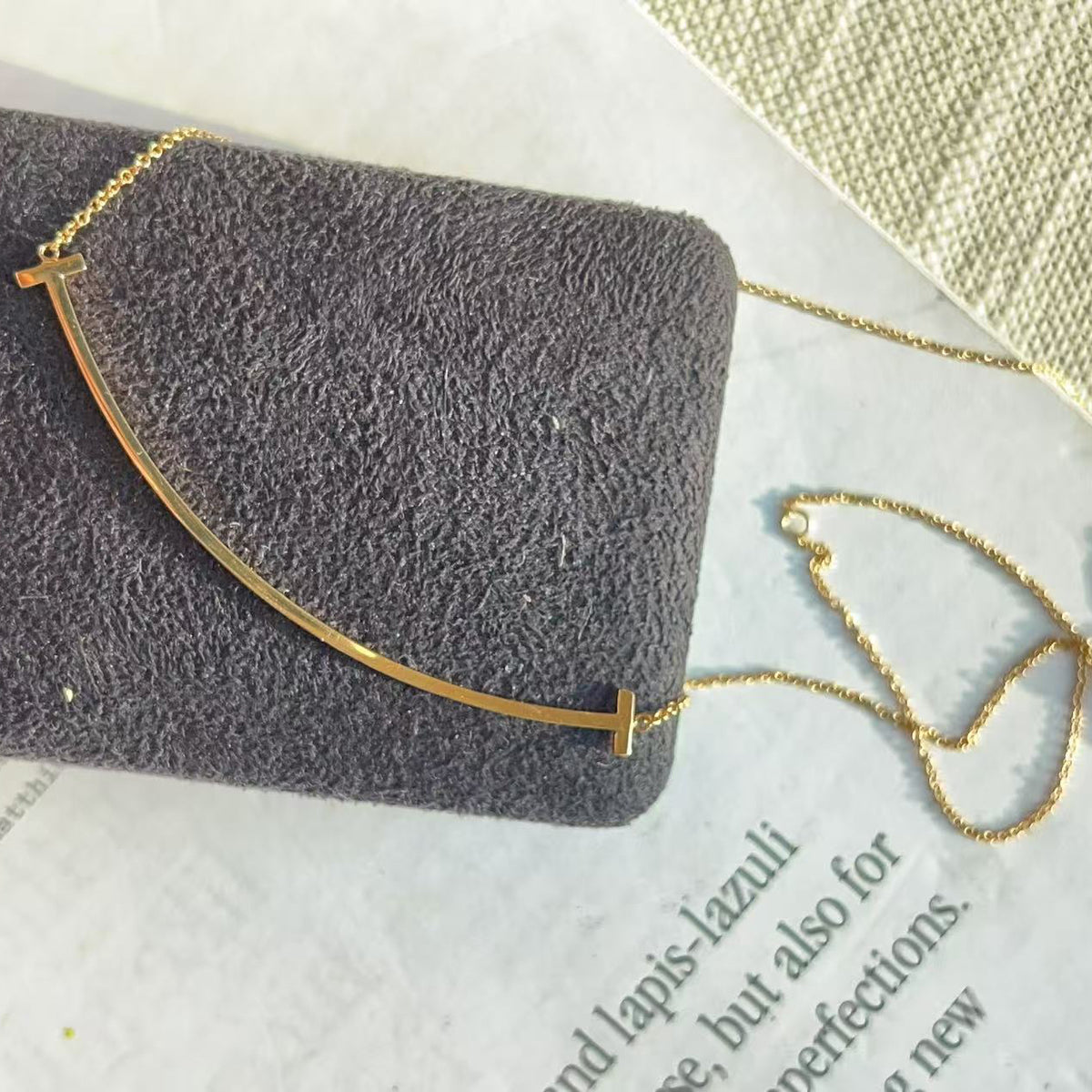 A gold-toned curved bar necklace displayed on a dark gray textured jewelry cushion, placed on a light surface with a woven textile in the background.