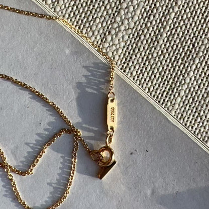 A close-up of a gold chain necklace with a small rectangular tag engraved with '14K 925', laid on a textured light gray surface next to a woven beige fabric.