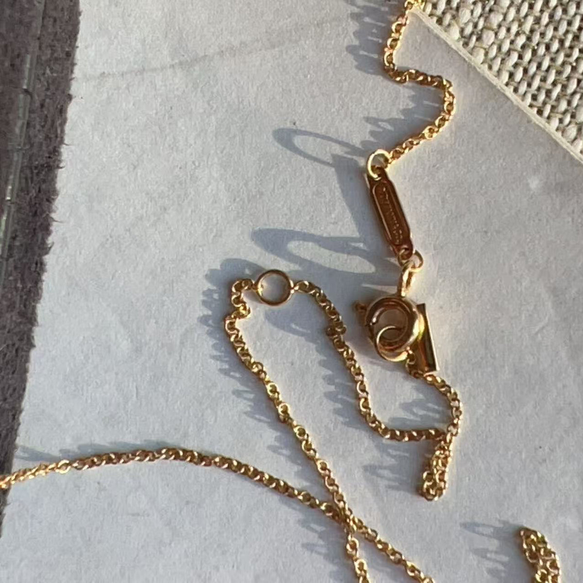 A gold chain necklace with a small rectangular pendant and lobster clasp, laid out on a light textured surface.