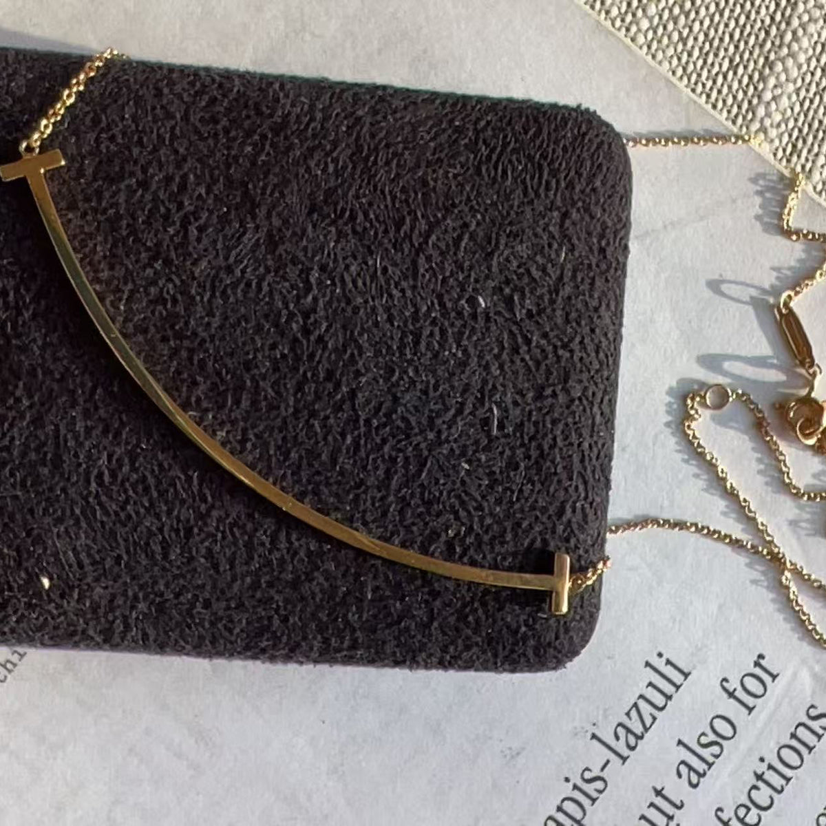 A gold-toned curved bar necklace with a cross pendant, displayed on a textured black velvet jewelry box over a light gray paper background.