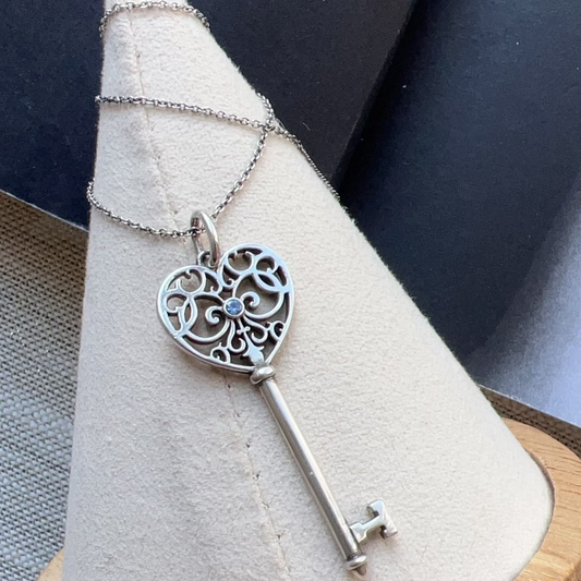 Silver key-shaped pendant on a heart-shaped stand with a blue gemstone.