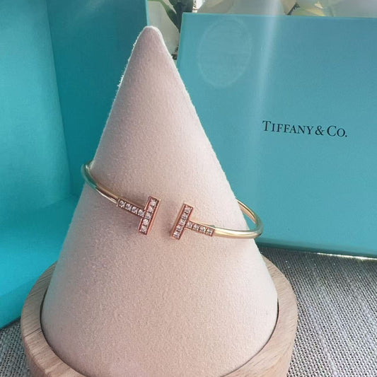 Rose gold letter 'T' bracelet on a pink stand with a Tiffany & Co. box in the background.