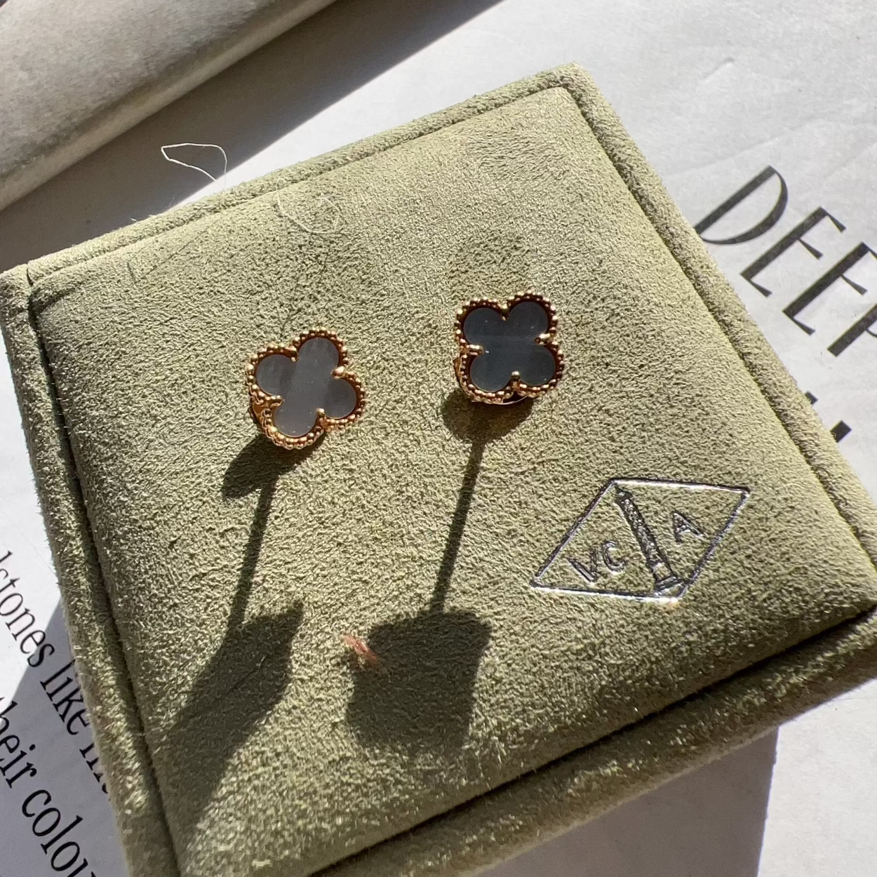 A pair of small clover-shaped stud earrings with gold-toned edges and dark gray centers, displayed on a textured olive-green jewelry box. The box has a silver-tone logo stamp and is photographed on a light-colored surface with strong sunlight and shadows.