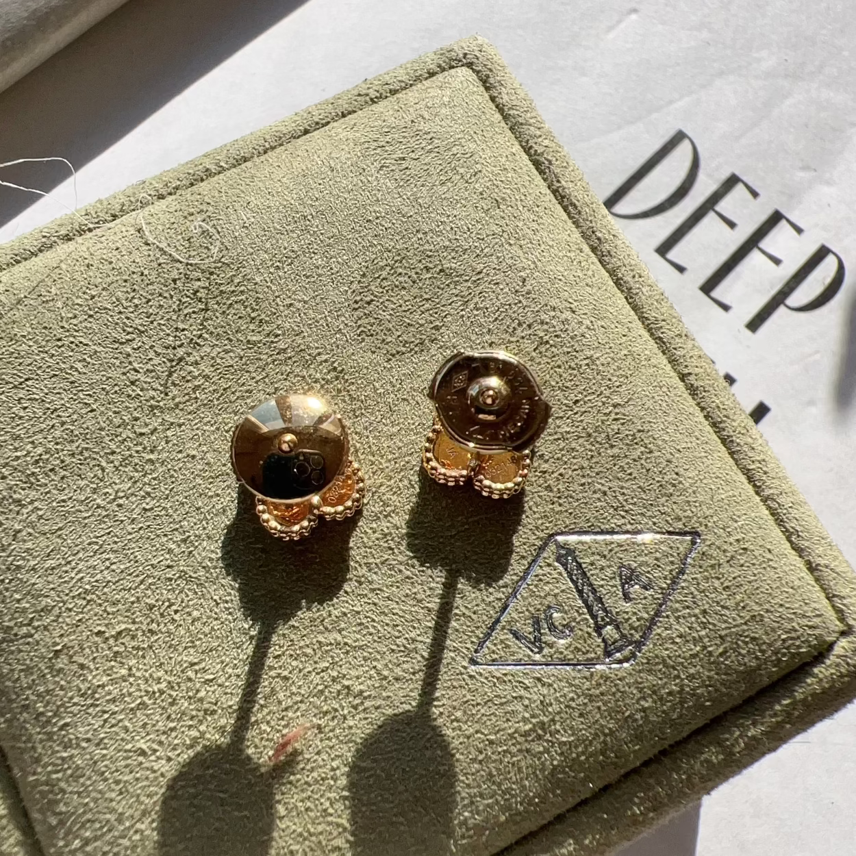 A pair of gold-toned stud earrings displayed on a textured olive-green jewelry box, with a white paper background showing the partial brand name 'DEEP'.