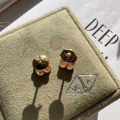 A pair of gold-toned stud earrings displayed on a textured olive-green jewelry box, with a white paper background showing the partial brand name 'DEEP'.