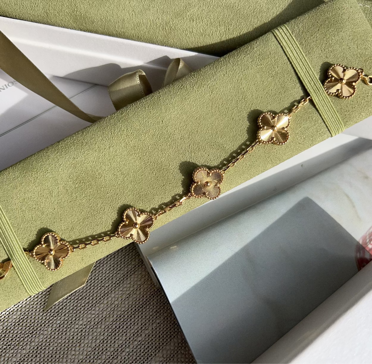 A gold-toned bracelet with four-link clover motifs laid diagonally across a light olive green velvet jewelry pouch, with white gift packaging and ribbon visible around it.