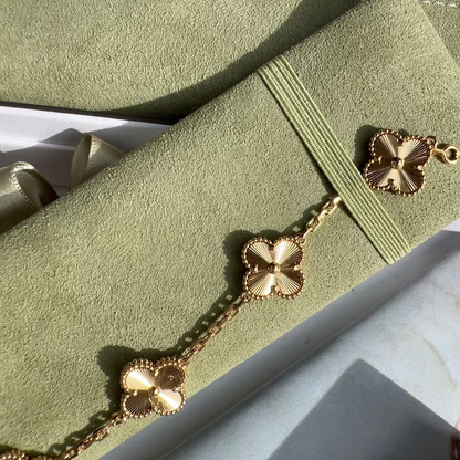 A gold-tone bracelet with four clover motifs laid diagonally across a sage green velvet pouch, with part of a white box visible in the background.