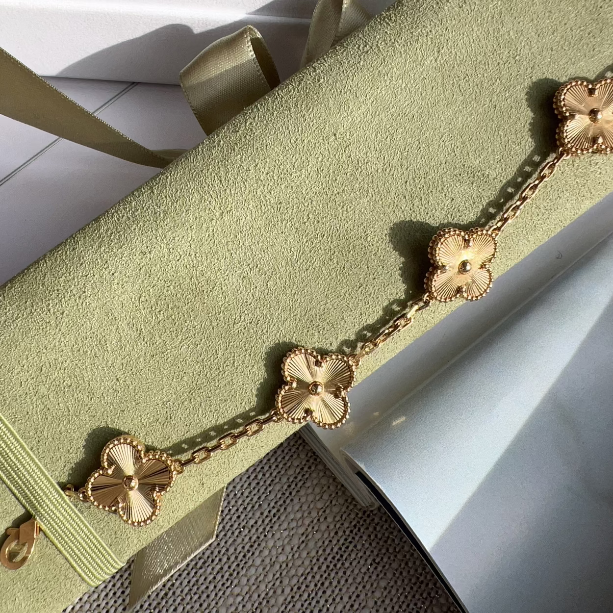 A gold-toned five-link chain bracelet with clover-shaped motifs, laid diagonally across a light sage green velvet jewelry box with a ribbon.