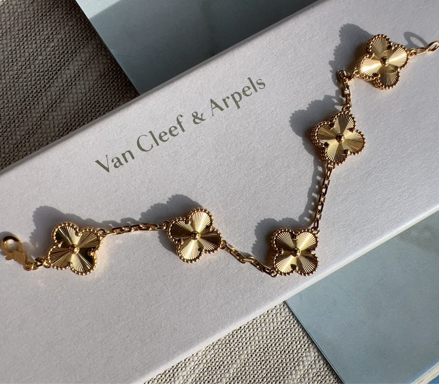 A gold-tone Van Cleef & Arpels Alhambra five-flower bracelet displayed on its white branded box, set against a neutral textured background.