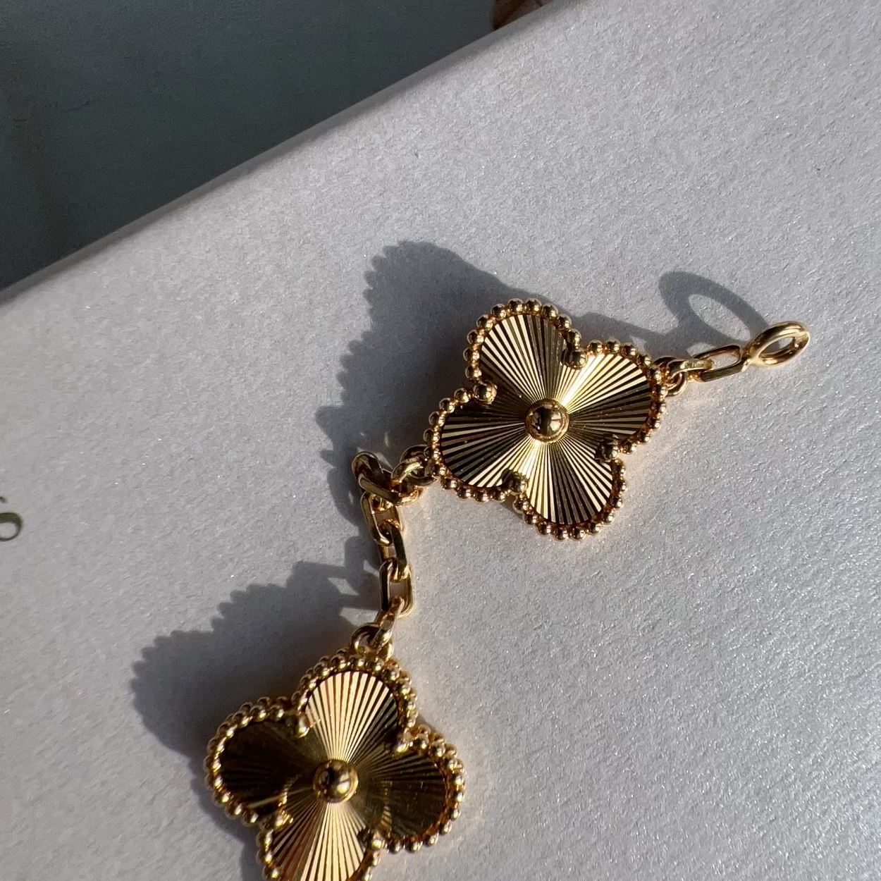 A pair of gold-toned clover-shaped earrings with textured, sunburst detailing and a short chain connector, resting on a white textured surface.