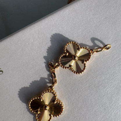 A pair of gold-toned clover-shaped earrings with textured, sunburst detailing and a short chain connector, resting on a white textured surface.