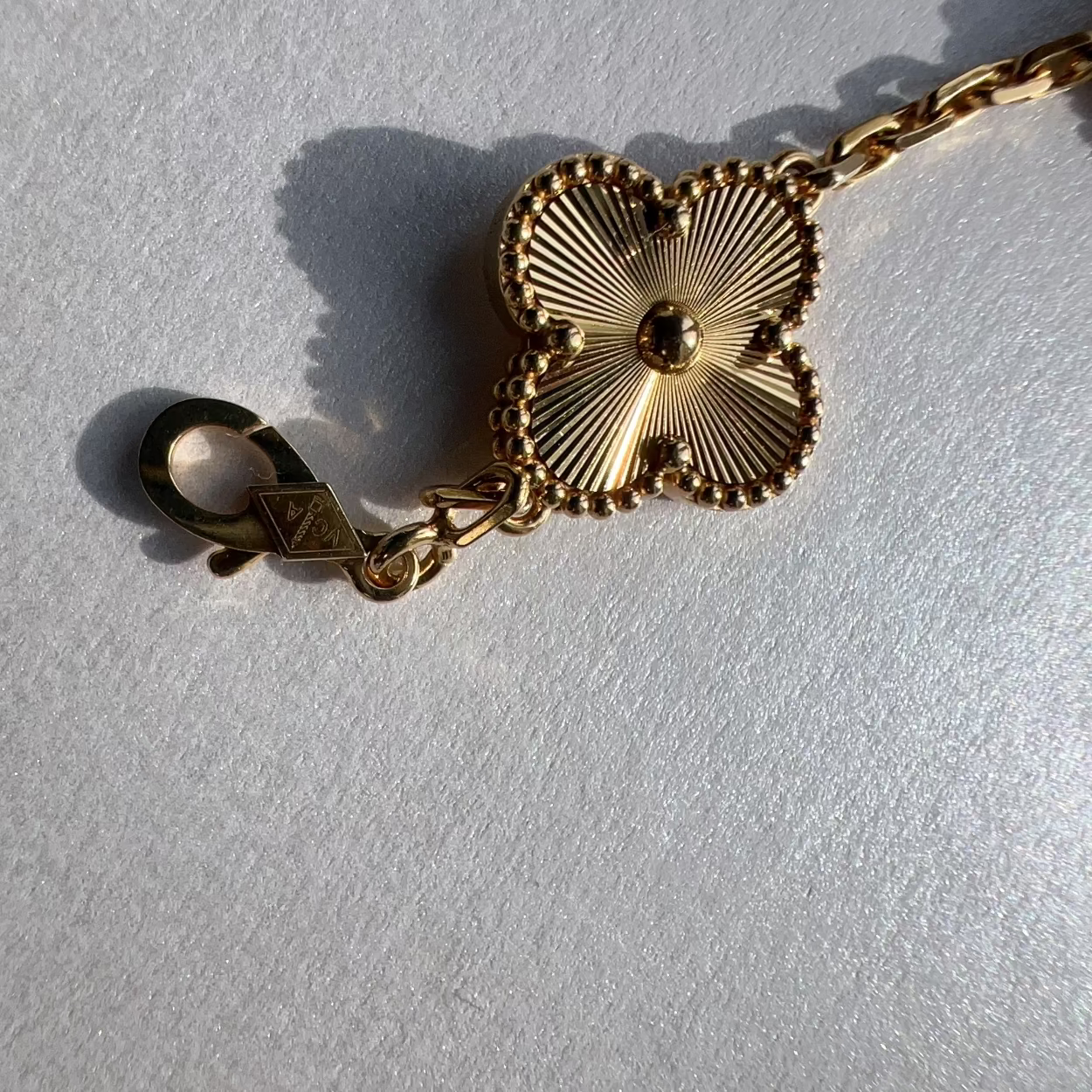 A gold-toned chain bracelet featuring a four-lobed clover or leaf-shaped charm with radiating petal details and a lobster clasp, photographed on a light textured surface.