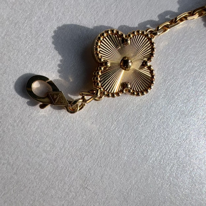 A gold-toned chain bracelet featuring a four-lobed clover or leaf-shaped charm with radiating petal details and a lobster clasp, photographed on a light textured surface.