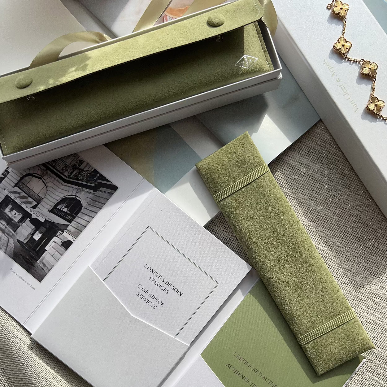 An olive green eyeglasses case and matching box are arranged on a light wood surface with printed cards and a gold-tone necklace nearby. No person or animal is present, so this is Product/Packaging rather than Lifestyle.