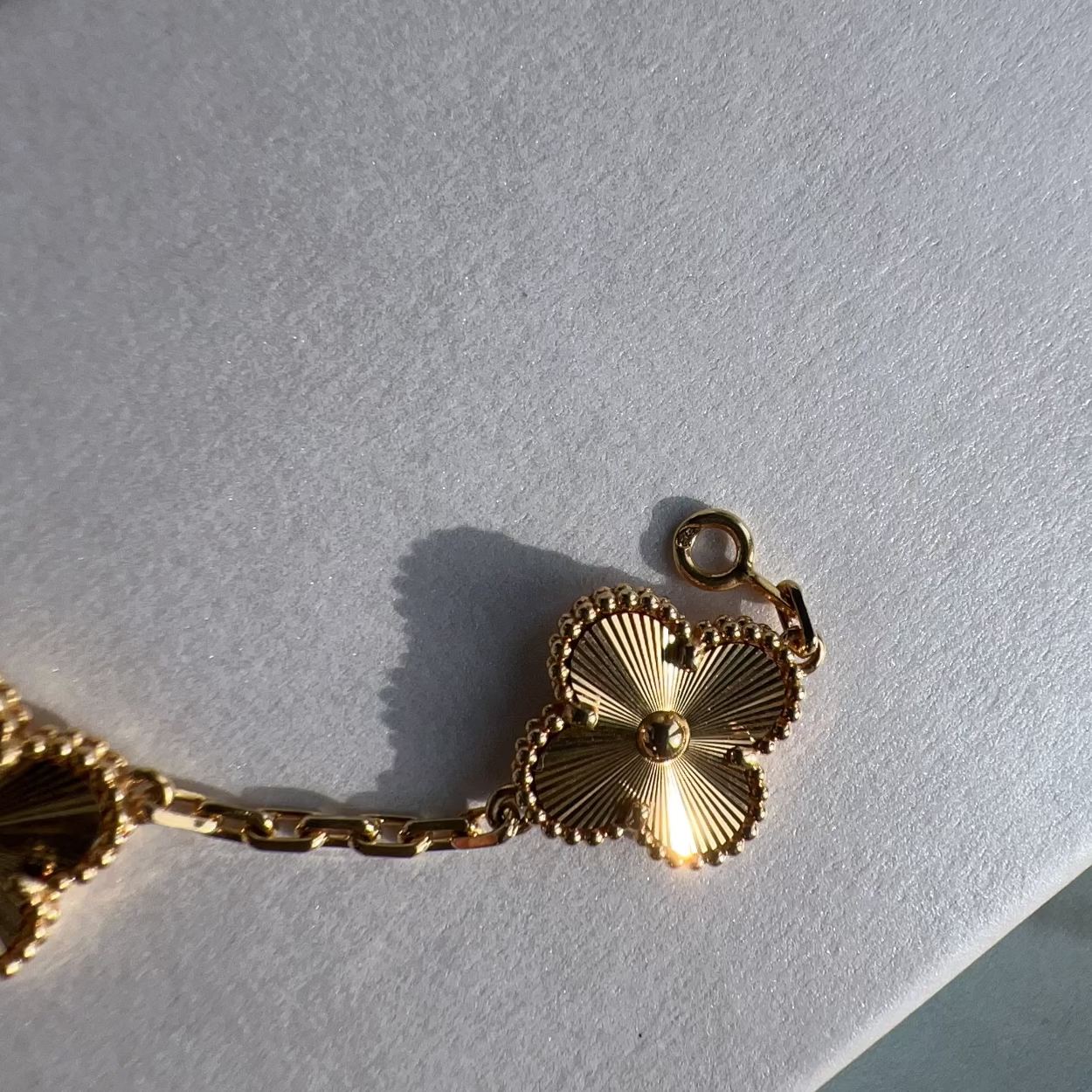 A close-up of a gold-tone bracelet with a textured, fan-like clover-shaped charm and matching chain links, resting on a white textured surface.