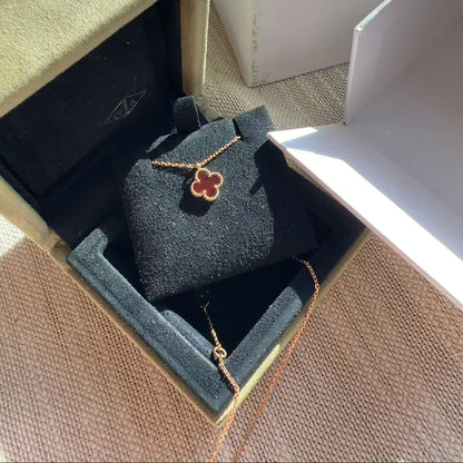A gold-toned necklace with a clover-shaped pendant is displayed inside an open jewelry box with a black velvet insert. A white Van Cleef & Arpels box is visible in the background on a textured beige surface.