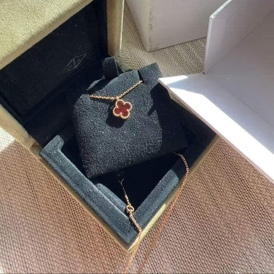 A gold-toned necklace with a clover-shaped pendant is displayed inside an open jewelry box with a black velvet insert. A white Van Cleef & Arpels box is visible in the background on a textured beige surface.