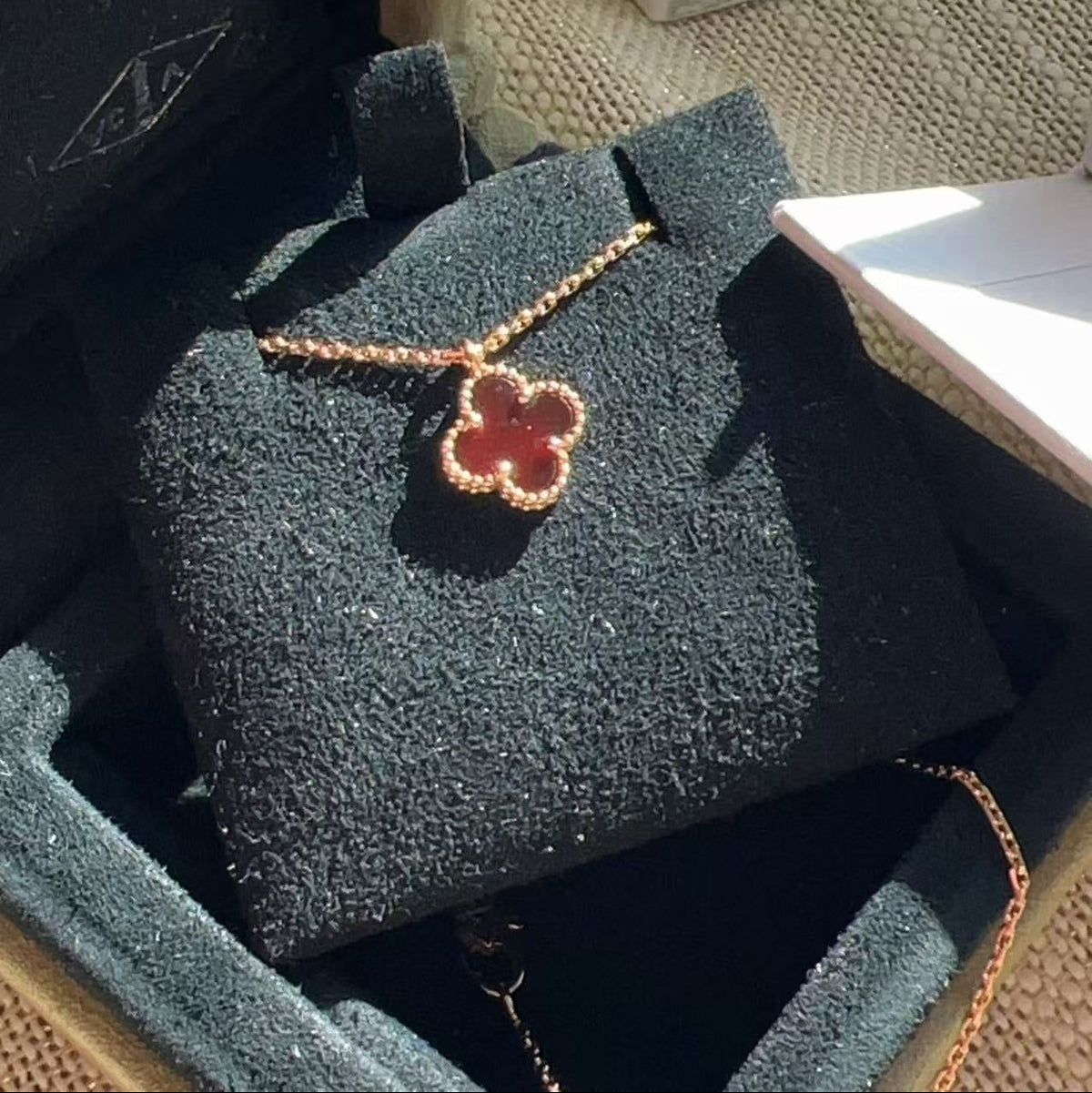 A rose gold four-leaf clover pendant necklace with a red center, displayed on a black velvet jewelry box inside an open presentation box.