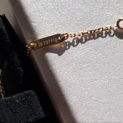 Close-up of a gold chain bracelet with a small rectangular tag engraved with 'JEW37115', resting on a textured white surface with a black velvet jewelry box visible at the side.