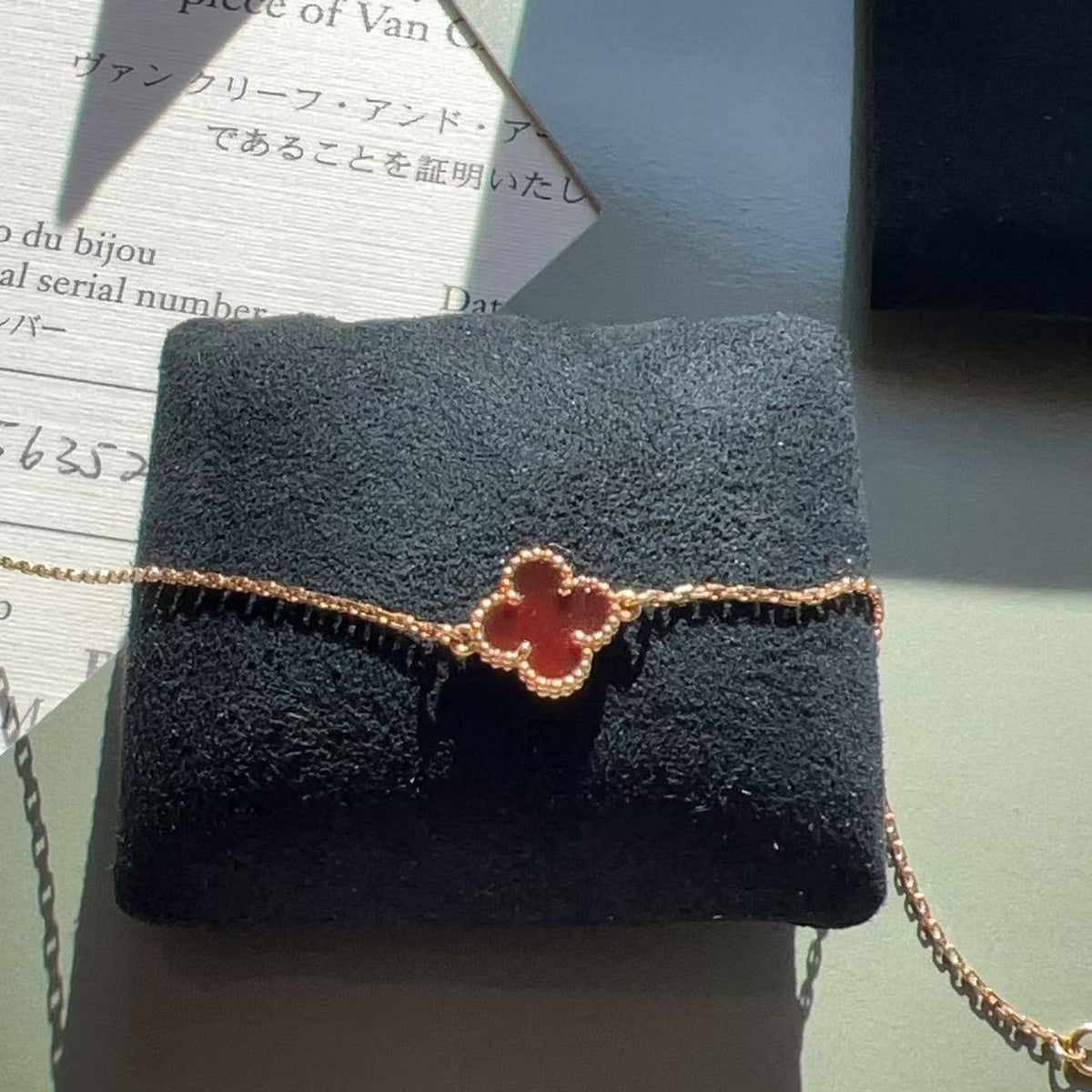 A delicate gold bracelet with a red clover motif, displayed on a black velvet jewelry cushion. The background features a light green surface with a Van Cleef & Arpels authenticity card and a black branded box partially visible.