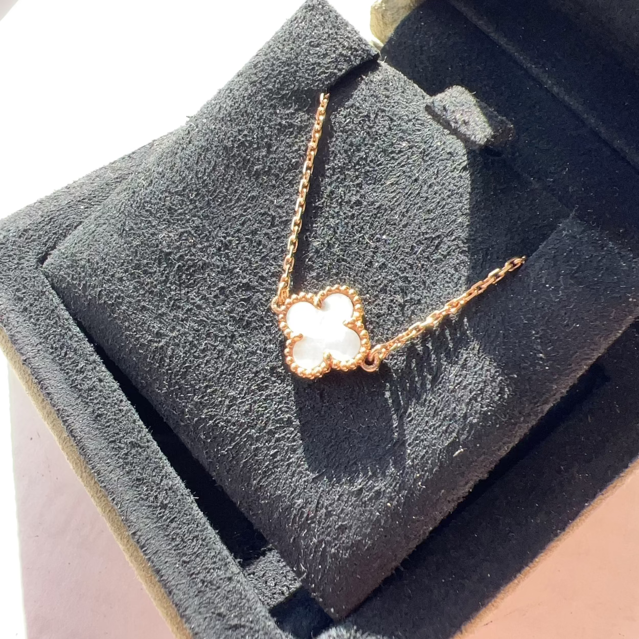A rose gold necklace with a clover-shaped pendant featuring a white inlay, displayed on a black velvet jewelry box.