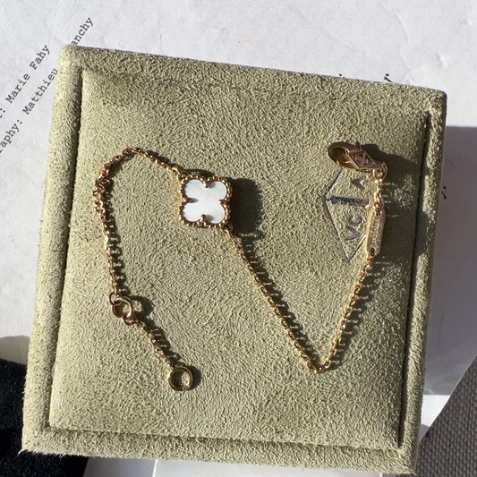 A gold-toned bracelet displayed on a textured beige jewelry box. The bracelet features a clover-shaped charm with a white inlay and a small heart-shaped charm with a gold anchor detail.