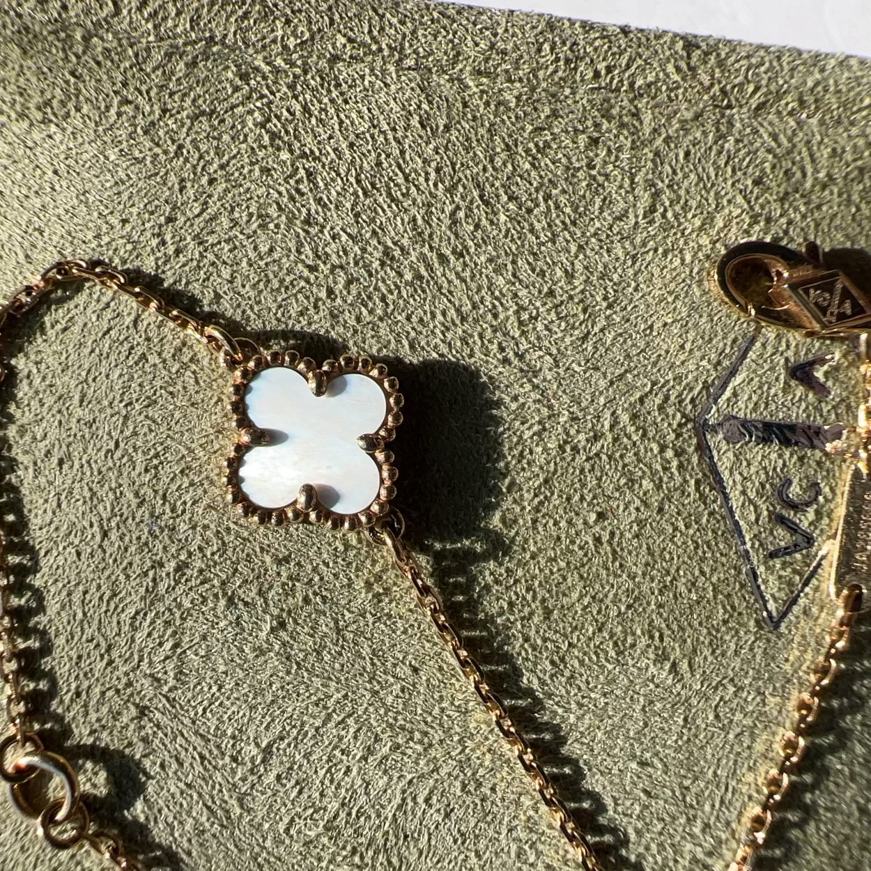 A gold-toned bracelet with a white mother-of-pearl four-leaf clover pendant, displayed on a textured olive-green jewelry box. A gold-toned VCA logo charm is visible on the chain.