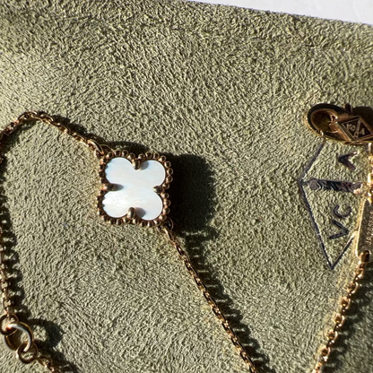 A gold-toned bracelet with a white mother-of-pearl four-leaf clover pendant, displayed on a textured olive-green jewelry box. A gold-toned VCA logo charm is visible on the chain.