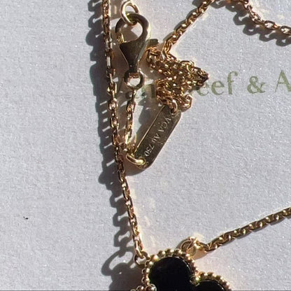Close-up of a gold chain necklace with a lobster clasp, tag, and part of a black clover pendant on a light background.