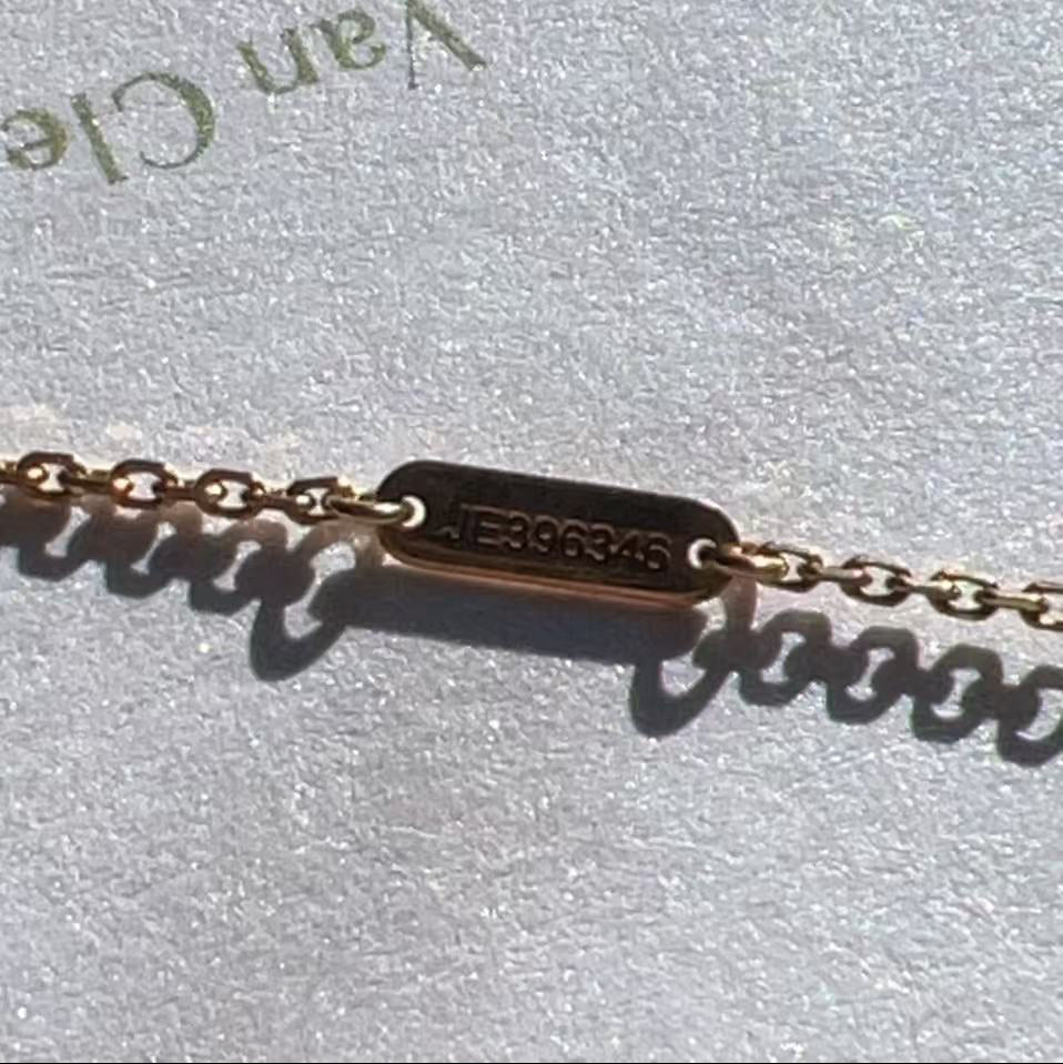 A close-up of a delicate gold chain bracelet with a rectangular engraved plate, resting on a textured light gray surface with partial 'Van Cle' text visible.